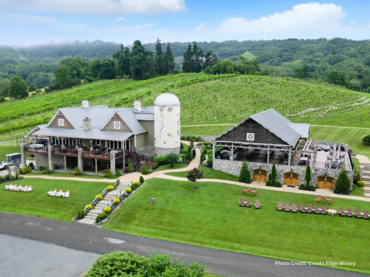 Aerial view of Creek's Edge Winery in Lovettsville, Virginia, showing the stone tasting room, barn, silo, and rolling vineyard rows on green hillside